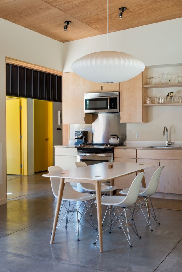 Bright open-plan kitchen and dining area with pale timber cabinetry, a matching timber-lined ceiling, polished concrete flooring, and white walls. The kitchen includes a compact stainless-steel range, microwave, sink, open shelving with glassware, and a dining table surrounded by molded white chairs. A large sculptural white pendant light hangs above the table, while a vivid yellow doorway provides a strong colour accent. Dark vertical screening is visible beside the doorway.