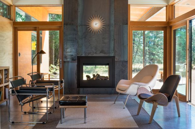 A warm, light-filled mid-century living room centred on a dark stone or concrete fireplace wall with a built-in firebox. Floor-to-ceiling timber-framed glazing surrounds the room and looks onto trees. The space contains iconic-style lounge chairs, a black leather office chair, a low ottoman, a glass-topped table, shelving, and a sculptural sunburst wall clock.