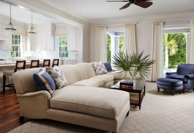 Bright, refined open-plan living and dining space with a cream upholstered sectional and chaise, patterned and navy accent cushions, a glass coffee table, blue armchair and ottoman, and a large neutral rug. The adjacent white kitchen features detailed paneled cabinetry, marble-look splashback surfaces, pendant lights, and a dining table. Tall curtained windows and French doors open to lush greenery, while a ceiling fan and traditional trim add architectural character.