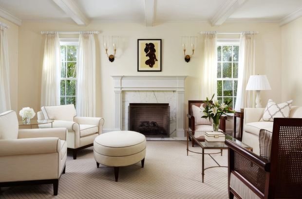 Bright, elegant living room with a symmetrical fireplace focal point, white painted mantel and surround, coffered ceiling beams, tall multi-pane windows with sheer curtains, cream upholstered seating, a round ottoman, glass-and-metal coffee table, and traditional decorative detailing. The room has a refined, warm neutral palette and abundant natural light.
