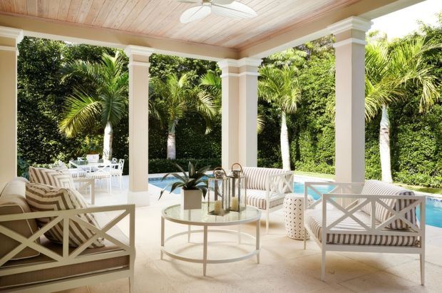 A bright, elegant covered outdoor living veranda with white classical columns and trim, a pale timber-panelled ceiling, light stone or concrete flooring, and white cross-back lounge furniture with striped cushions. The open-sided space overlooks a turquoise swimming pool and dense tropical planting, creating a refined resort-like setting.