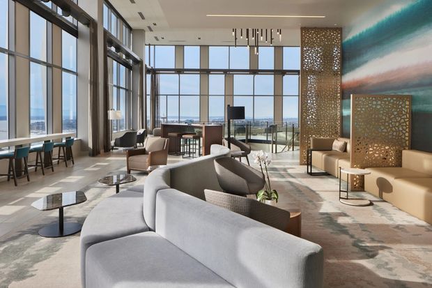 A spacious, high-end contemporary commercial lounge or hotel lobby with floor-to-ceiling glazing, expansive city and water views, sculptural grey seating, small geometric coffee tables, lounge chairs, and a long built-in bar-height counter. Decorative perforated metal screens divide seating zones, while a large abstract turquoise, white, and terracotta wall feature adds colour. The space has a high ceiling, polished stone-like flooring, abundant natural light, and refined modern furnishings.