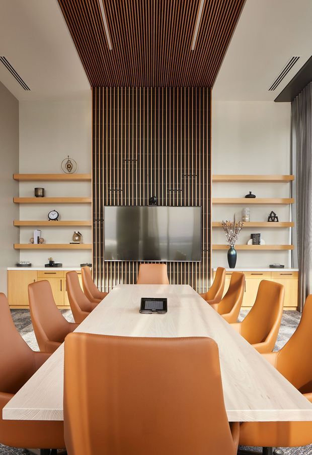 A contemporary dining and meeting room centered on a long pale wood table with numerous caramel leather chairs. The rear wall features extensive light wood built-in cabinetry and open display shelving, with a dark vertical timber-slat feature wall and matching slatted ceiling framing a wall-mounted television. Neutral plaster walls, concealed linear air vents, curtains, and soft recessed lighting complete the refined, warm interior.