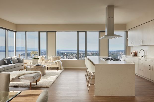 Open-plan contemporary apartment interior combining a bright kitchen with a large island and integrated rangehood, dining seating, and a living area. Floor-to-ceiling glazing provides expansive views across a coastal city and water, while pale cabinetry, light flooring, neutral upholstery, and warm wood accents create a refined, airy setting.
