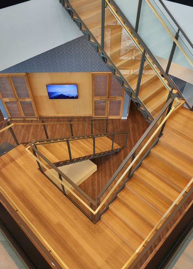 A dramatic top-down view of a multi-level interior stairwell with warm timber stairs, landings and flooring, framed by dark metal railings and clear glass balustrades. The stair flights connect around a tall open void, with timber wall panelling, built-in storage, a wall-mounted television and a patterned blue-grey feature wall visible below. The composition emphasises the geometric circulation and double-height volume.