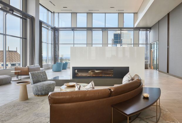 A spacious contemporary living room or lounge with floor-to-ceiling glazing, expansive urban views, pale timber flooring, and a large suspended or integrated fireplace set into a white feature wall. A curved brown leather sectional sofa, sculptural lounge chairs, and a dark timber coffee table form a refined seating arrangement around the fireplace. The room has a high ceiling, abundant natural light, and a polished apartment or commercial hospitality character.