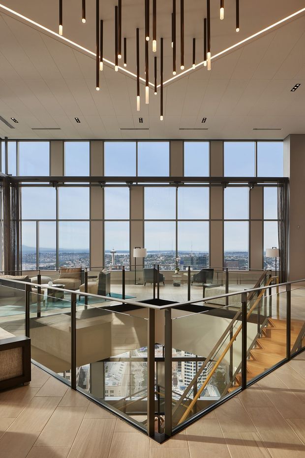 A refined contemporary multi-level interior with a large glazed atrium or stair void overlooking a dense city skyline. The space features floor-to-ceiling windows, slim dark metal-framed glass balustrades, pale large-format tiled flooring, timber stairs with a dark handrail, and a striking ceiling installation of numerous suspended linear pendant lights. Lounge seating and tables are arranged around the perimeter, with abundant daylight and expansive urban views.