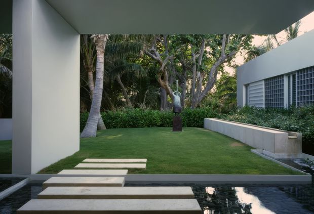 A landscaped courtyard garden viewed from a modern residence, with rectangular stepping stones crossing a narrow reflective water feature toward a manicured lawn. White rendered walls, a low raised planter, dense tropical planting, and a sculptural artwork create a calm contemporary outdoor composition.