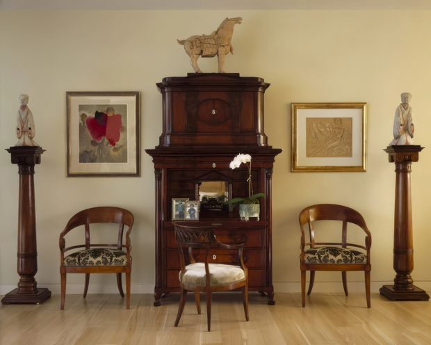 A formally arranged traditional interior vignette featuring a tall, dark timber antique display cabinet or secretary desk centered against a warm cream wall. Matching dark timber pedestal columns with figurative sculptures flank the cabinet, while two upholstered antique armchairs sit symmetrically on either side. Framed artwork, a horse sculpture, decorative objects, and a small orchid complete the composition. The room has pale timber flooring and a warm, refined, museum-like atmosphere.