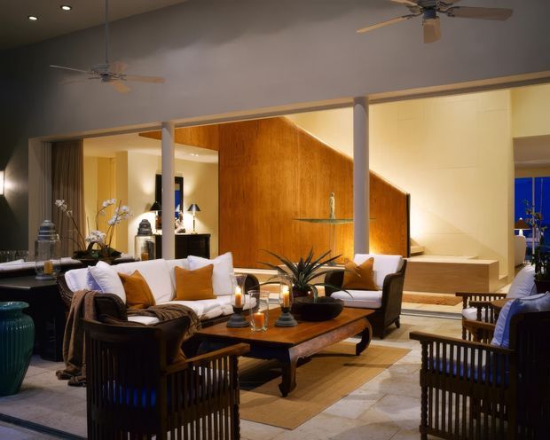 A spacious, warmly lit open-plan living room with a sunken seating area, white sofas, dark wood lounge chairs, and a large wooden coffee table. The room features a dramatic angled timber-clad feature wall, exposed columns, recessed ceiling lighting, ceiling fans, tiled flooring, and steps leading to an elevated adjacent area.
