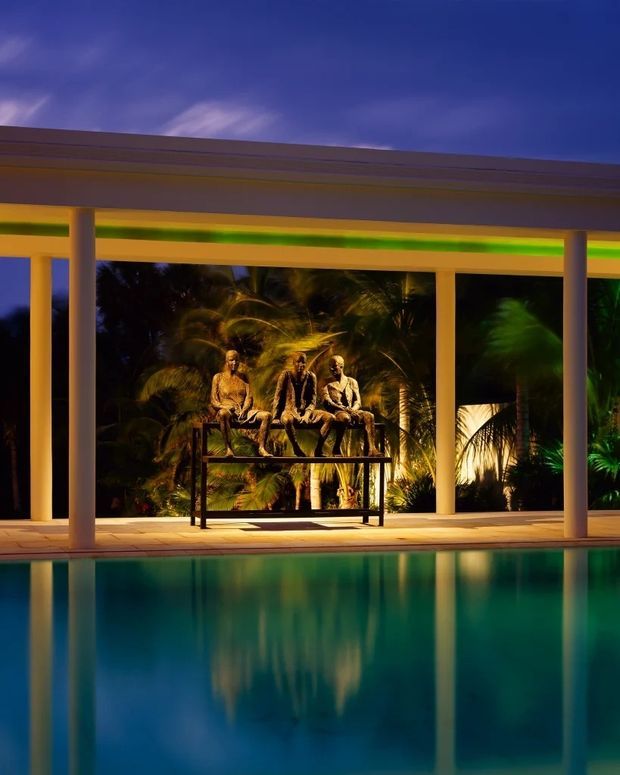 Night-time view of a modern covered pool pavilion with slender square columns, a broad flat roof, and warm architectural lighting. A tranquil swimming pool reflects the structure, while tropical planting and three sculptural figures form a dramatic backdrop.