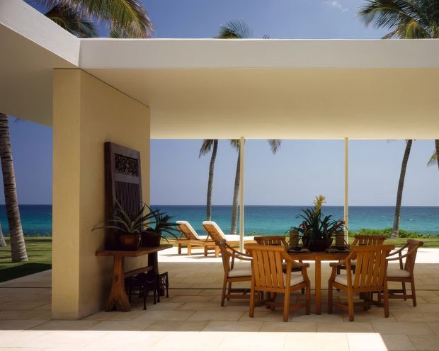 A covered outdoor dining terrace overlooking a turquoise ocean, with a large flat roof canopy, pale stucco columns and walls, stone or tiled flooring, and timber dining furniture. Tropical palms and potted plants frame the open view toward the beach and sea.