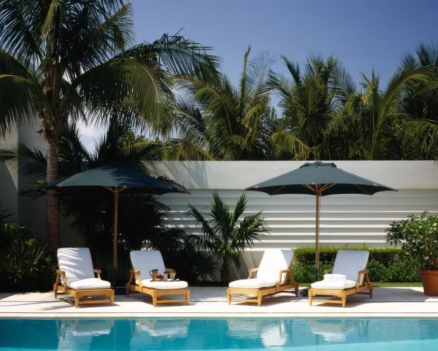 Bright tropical poolside terrace with a turquoise swimming pool, white rendered privacy wall featuring horizontal relief bands, lush palms and greenery, and multiple timber-framed white-cushioned sun loungers beneath dark umbrellas. The scene has a relaxed resort-like outdoor character.