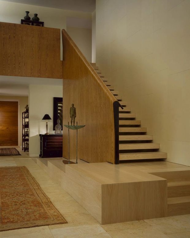 A refined interior stair hall with a floating timber staircase enclosed by a tall timber screen and integrated stepped platform. Pale stone or limestone flooring and wall surfaces create a warm, neutral backdrop, with dark timber furniture, rugs, lamps, and sculptural objects visible in the adjoining entry and living areas. The composition is architectural and minimalist, with strong clean lines and a high ceiling.
