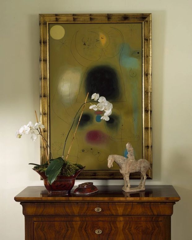 A warmly lit interior vignette featuring a dark polished timber console cabinet with ornate veneer, drawers, and brass key escutcheons. A large framed abstract artwork hangs above it, accompanied by a potted white orchid, a small decorative vessel, and a carved horse sculpture. The composition has a refined, traditional character with rich wood tones and muted olive, cream, and black accents.