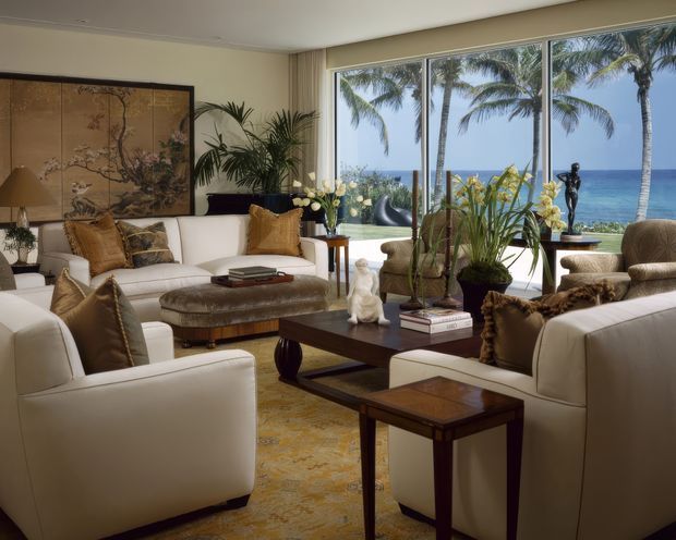 A refined living room overlooking a tropical ocean through expansive floor-to-ceiling windows. The room features cream upholstered sofas and armchairs, dark wood coffee and side tables, a patterned area rug, potted tropical plants, decorative orchids, a large framed botanical artwork or screen, and layered table and floor lighting. Palm trees and the sea are visible beyond the glazing.