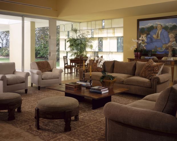 A spacious, warm-toned mid-century living room with extensive glazing, a patterned carpet, upholstered sofas and armchairs, carved wood coffee table, decorative stools, indoor plants, and a dining area beyond. A large colourful artwork forms a focal point above the timber sideboard, while translucent geometric glazing and simple built-in architectural lines add character.