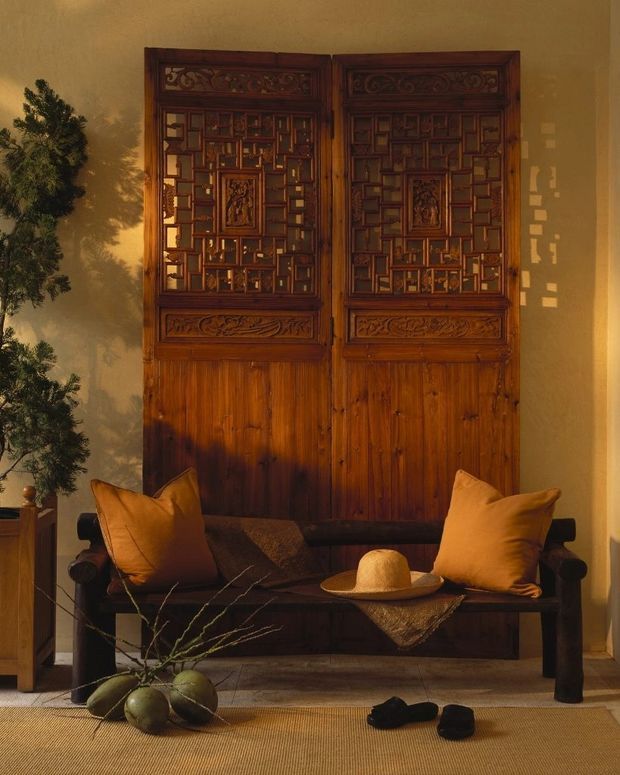 A warmly lit interior vignette featuring an ornate pair of dark timber carved doors or screens with intricate geometric and floral detailing. A dark wooden bench sits in front, styled with ochre cushions, a woven throw, a straw hat, and natural branches with green fruit. The space has a rich, earthy palette and traditional handcrafted character.