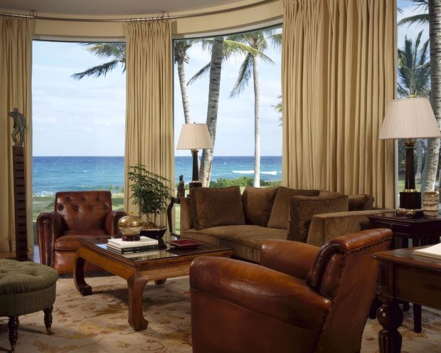 A refined living room with expansive floor-to-ceiling glazing overlooking a tropical beachfront and ocean. The room features rich brown leather armchairs and sofa seating, a substantial carved wood coffee table, traditional table lamps, patterned curtains, and a neutral patterned rug. The composition is warm and classic, with palm trees and the sea visible beyond the windows.