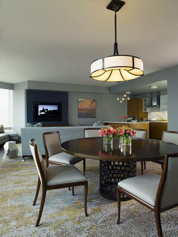 A contemporary open-plan dining and living area with a round dark dining table, upholstered wooden dining chairs, patterned area rug, and a prominent circular pendant light. A grey sofa sits behind the dining setting, with a dark media wall and artwork nearby. The open kitchen is visible in the background, featuring warm timber cabinetry, pale benchtops, and pendant lighting. The room has muted grey walls and a refined, coordinated palette.