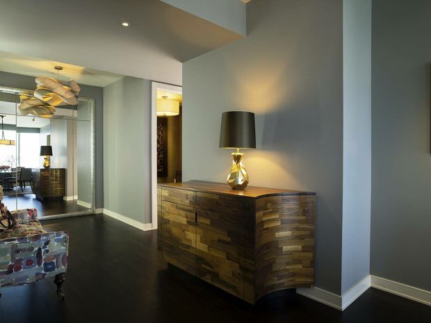 A refined contemporary interior hallway or entry area with dark flooring, cool grey walls, white trim, and a sculptural timber sideboard topped with a decorative lamp. A large mirror reflects an adjacent dining area with pendant lighting and patterned upholstered seating. The space has layered lighting, custom-looking joinery, and a sophisticated, moody atmosphere.