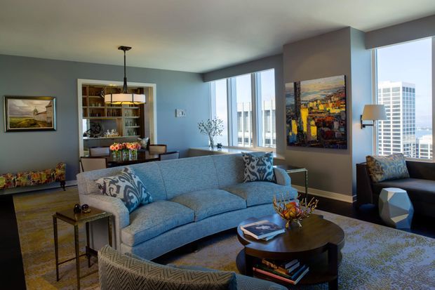 A refined high-rise living room with a large blue upholstered sofa, patterned accent cushions, dark wood round coffee table, armchairs, and a sculptural pendant light. Built-in shelving and a small dining area sit at the rear, while expansive windows provide an urban city view. The room combines cool blue-grey walls and upholstery with warm timber flooring, colourful artwork, and layered decorative furnishings.