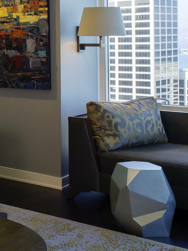 A contemporary high-rise interior corner featuring a dark upholstered armchair or compact sofa, a sculptural faceted side table, patterned accent cushion, wall-mounted lamp, abstract artwork, and large windows overlooking surrounding city buildings and water. The space has dark flooring, neutral walls, and strong geometric and colourful decorative accents.