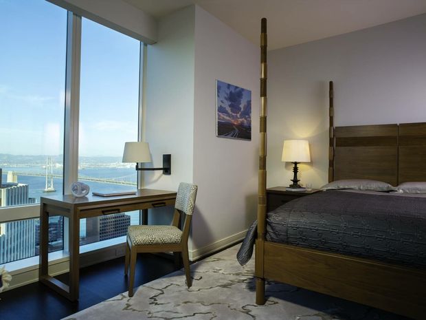A compact, well-appointed bedroom in a high-rise setting, featuring a dark wood platform bed with a tall paneled headboard, matching dark wood writing desk, upholstered desk chair, bedside lamps, and a patterned area rug. Full-height glazing provides expansive waterfront and city views. A distinctive vertical wooden decorative screen or post separates the sleeping area from the desk zone.