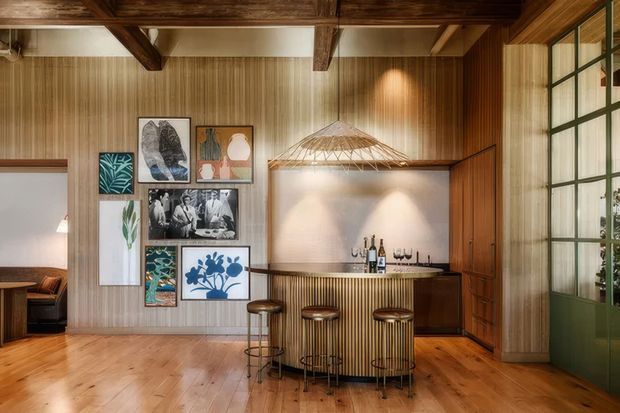 Warm, richly detailed home bar or entertaining area with a curved fluted timber bar, three stools, bottle storage, and a large woven pendant light. The room features honey-toned timber flooring, vertical timber wall panelling, exposed dark timber ceiling beams, framed artwork, and dark green framed glazing or a partition on the right. A doorway opens to an adjacent room on the left.