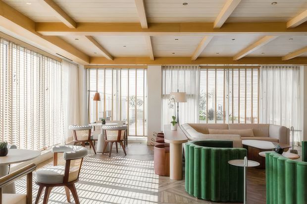 Bright, spacious open-plan living and dining room with extensive floor-to-ceiling glazing fitted with vertical blinds, a warm timber-beamed and panelled ceiling, and patterned timber flooring. The room includes a dining table and chairs, a curved neutral sofa, sculptural green upholstered lounge seating, side tables, and indoor plants. Strong natural light creates a refined contemporary atmosphere.
