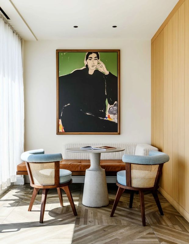 A refined, light-filled sitting and dining nook with two mid-century-inspired cane armchairs arranged around a small sculptural pedestal table. A curved upholstered banquette sits against the rear wall beneath a large framed portrait artwork, while full-height sheer curtains line the left side and warm vertical timber wall panelling defines the right side. The floor features a geometric herringbone timber pattern, creating a sophisticated, warm contemporary interior.