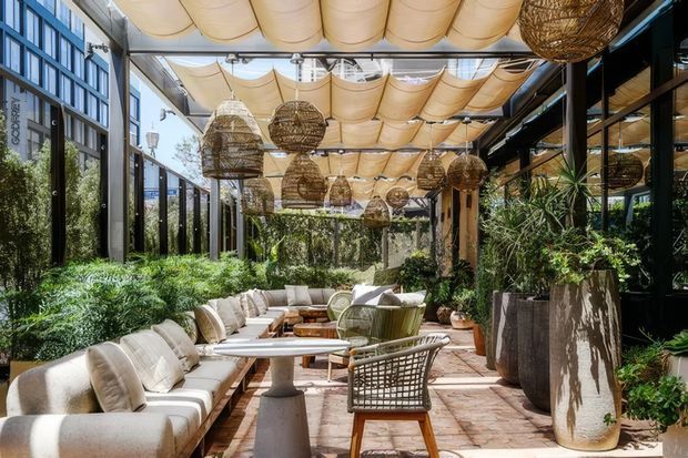 A bright, enclosed outdoor courtyard lounge with floor-to-ceiling glazing, abundant greenery, cushioned seating, café-style tables, and woven pendant lights. A translucent fabric canopy creates a warm, dappled ceiling, while terracotta-toned paving and natural woven furnishings give the space a relaxed resort-like atmosphere.