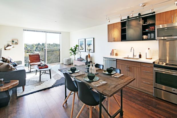 Open-plan contemporary apartment interior combining a compact kitchen and dining area with a living room. The kitchen features warm wood cabinetry, a white benchtop, stainless-steel appliances, open black shelving, and track lighting. A timber dining table with dark chairs sits in the foreground, while the living area has a grey sofa, coffee table, rug, and large glazed doors overlooking an urban landscape.