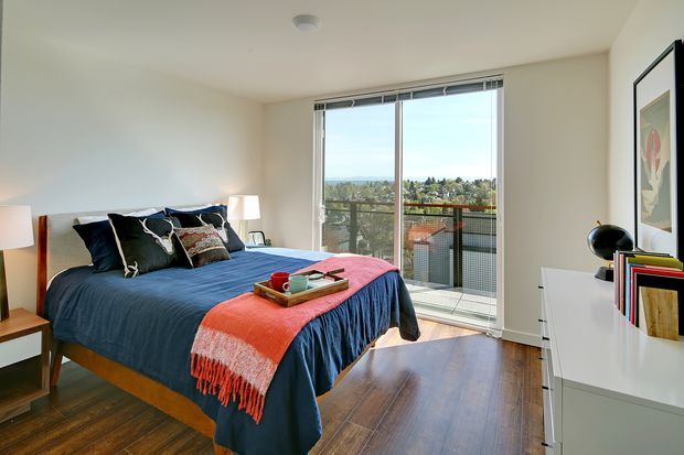 A bright, contemporary bedroom with a neatly made bed dressed in deep blue bedding and a coral throw, set against white walls. A large sliding glass door opens to a balcony with views over neighbouring rooftops and trees. The room includes timber flooring, white bedside furniture, a white dresser, black bedside and tabletop lamps, and framed wall art.