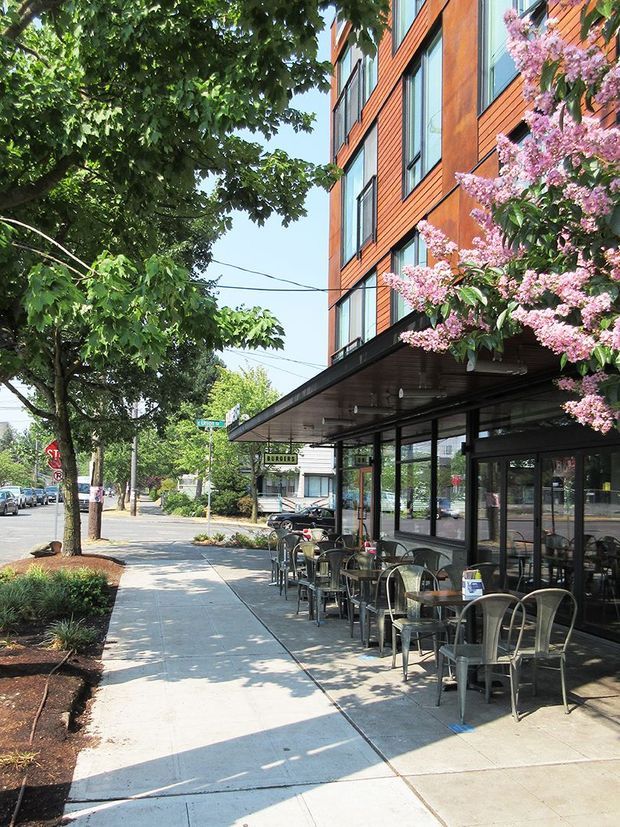 Street-level exterior view of a contemporary multi-storey commercial building with warm timber-clad upper floors, extensive dark-framed glazing, and a deep projecting canopy. Outdoor café seating occupies the wide concrete sidewalk, with mature street trees and flowering planting framing the urban frontage.