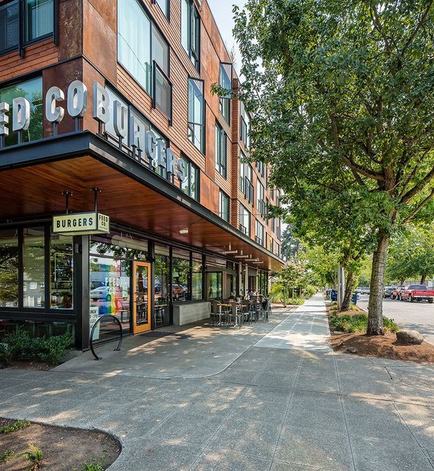 Street-level view of a contemporary multi-storey mixed-use building with ground-floor restaurant and retail spaces, upper-level timber-clad facade, large windows, projecting signage, outdoor café seating, and a tree-lined urban sidewalk.