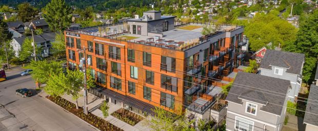 Aerial view of a contemporary multi-storey apartment building with a bold orange-brown brick facade, extensive dark-framed glazing, projecting balconies, and a flat rooftop terrace. The building is situated in a leafy urban or suburban neighbourhood alongside detached houses and tree-covered hills.