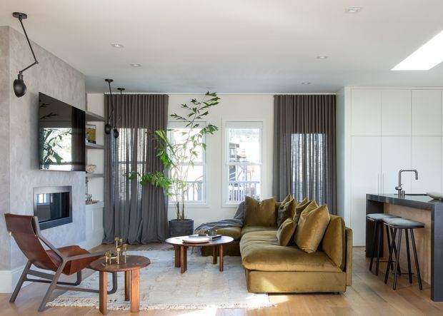 A refined open-plan living room with a warm ochre sectional sofa, mid-century-style leather lounge chair, nested timber coffee tables, and a concrete or stone fireplace wall with wall-mounted television. Full-height grey curtains frame the windows, while a compact kitchen with dark cabinetry and a peninsula is visible at right. The room has light timber flooring, white walls and ceiling, recessed lighting, and a skylight, creating a bright contemporary interior with warm accents.