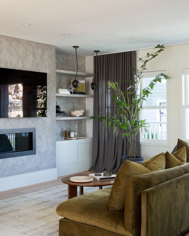 A refined contemporary living room with a wall-mounted television and fireplace set into a textured grey feature wall. Built-in open shelving and white lower cabinetry sit beside dark full-height curtains and a large indoor plant. A plush olive-brown velvet sofa, round timber coffee table, and pale textured rug create a warm, layered seating area. Windows bring in daylight and show a small exterior balcony or neighbouring structure.