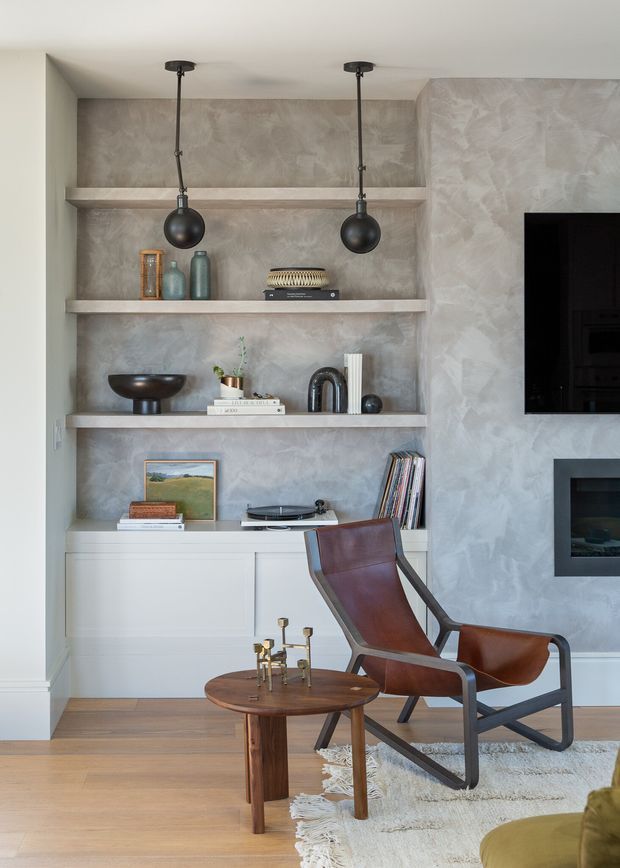 A refined living room featuring a built-in media and display wall with open shelves, textured plaster finish, white lower cabinetry, and a wall-mounted television with fireplace below. The room includes a brown leather lounge chair, round timber side table, neutral rug, decorative objects, books, record player, and black pendant lights. The palette is warm and neutral with mid-century-inspired furnishings.