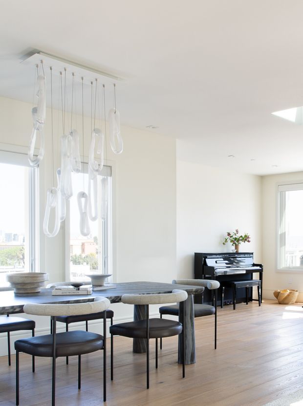 Bright, high-ceilinged open-plan dining and living space with large windows, pale timber flooring, a dark stone dining table surrounded by upholstered dining chairs, and a black upright piano against the far wall. A sculptural cluster of suspended translucent pendant lights hangs above the table. The interior is predominantly white and softly illuminated by daylight, with a minimal, refined aesthetic.
