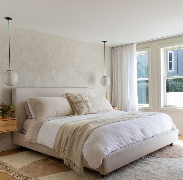 A softly styled bedroom with a low upholstered bed, layered white and beige bedding, textured accent cushions, and a warm neutral palette. The room features a sculptural textured feature wall, sheer full-height curtains, large white-framed windows, minimalist globe pendant lights, and a timber bedside table. Natural daylight creates a calm, inviting atmosphere.