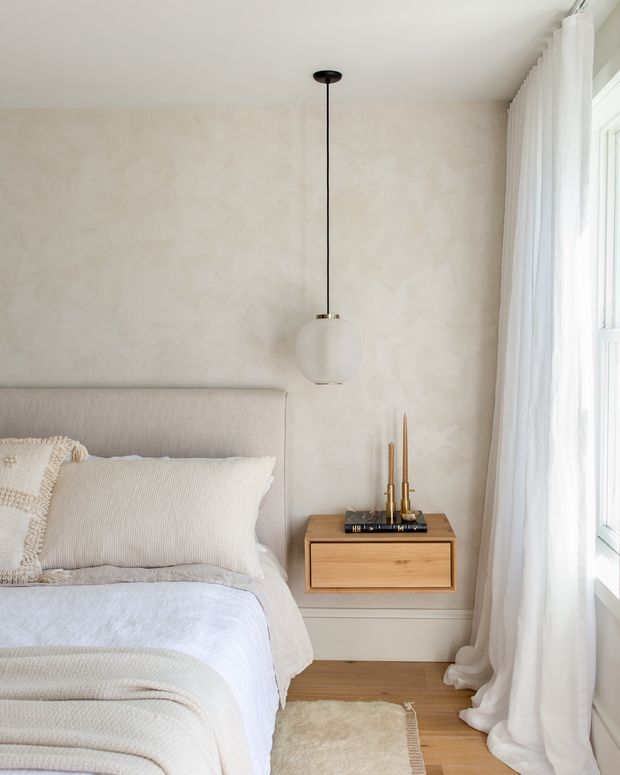 A softly styled bedroom corner with a neutral upholstered bed, layered cream and white bedding, a floating light-wood bedside cabinet, brass candlesticks, and a simple white globe pendant. The room has a textured plaster-like beige wall, pale timber flooring, and full-height sheer white curtains beside a bright window. The overall look is calm, warm, refined, and minimalist.