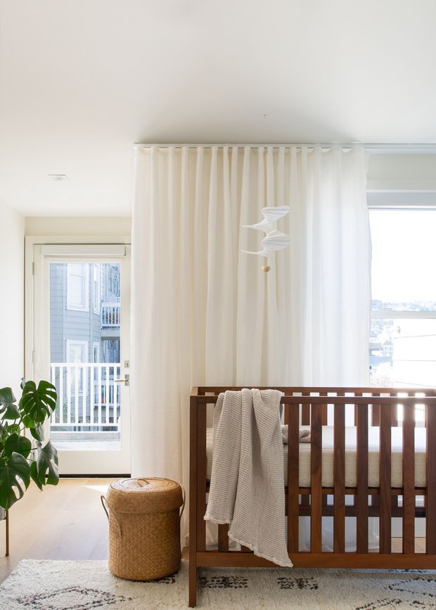 Bright, calm nursery or bedroom corner with a dark timber cot in the foreground, cream floor-to-ceiling curtains, pale flooring, a woven basket, soft rug, leafy plant, and a sculptural hanging mobile. A glazed door and window bring in abundant daylight and show an exterior balcony beyond.
