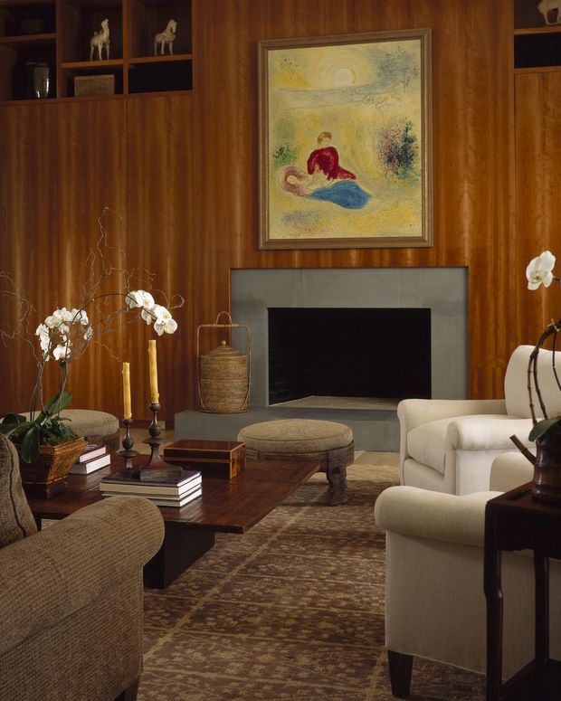 A warm, richly furnished living room centred on a fireplace with a grey tiled surround, framed artwork above, and extensive dark timber wall panelling and built-in shelving. Upholstered armchairs and a sofa surround a dark timber coffee table, with patterned rug, books, candles, and orchid arrangements adding traditional decorative detail.