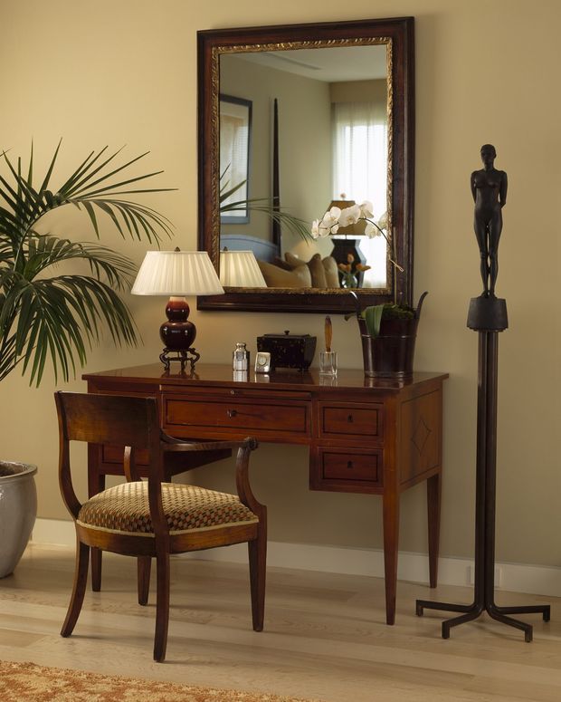 A refined interior vignette featuring a traditional dark timber writing desk with drawers, an upholstered wooden chair, table lamp, large ornate framed mirror, potted plants, and a slender sculptural floor lamp. The warm neutral wall and pale timber flooring create an elegant, softly lit entry or study-like setting.