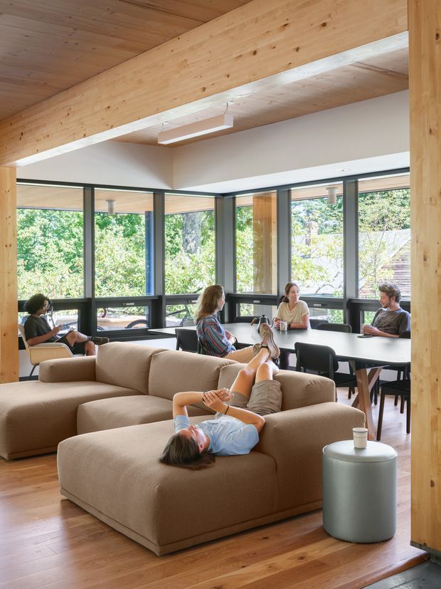 Open-plan living and dining space with exposed timber beams and ceiling, extensive dark-framed corner glazing, warm timber flooring, a large taupe sectional sofa, and a dining table surrounded by people. The room is bright and informal, with views of leafy surroundings through the windows.
