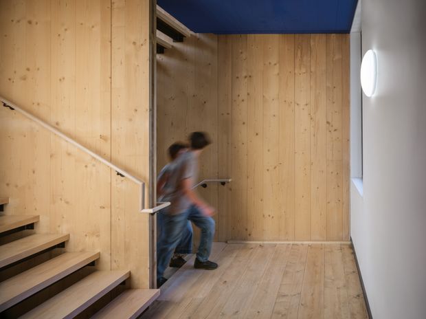 Interior view of a timber-lined stair hall or landing. Exposed light wood wall panelling, matching timber flooring, open-riser stairs with simple metal handrails, and a deep blue ceiling panel create a warm yet contemporary architectural interior. A blurred person is moving through the space, and a round wall light is mounted on the white wall at right.