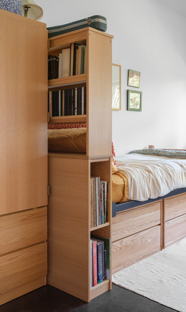 A bright, compact bedroom featuring extensive built-in light timber joinery. A tall open bookshelf separates a wardrobe from a raised bed with integrated timber drawers beneath. The bed is dressed in a striped cream and white duvet with patterned cushions, while framed artwork hangs on the white wall. A dark floor and pale rug provide contrast, with a vintage suitcase and decorative objects stored above the shelving.