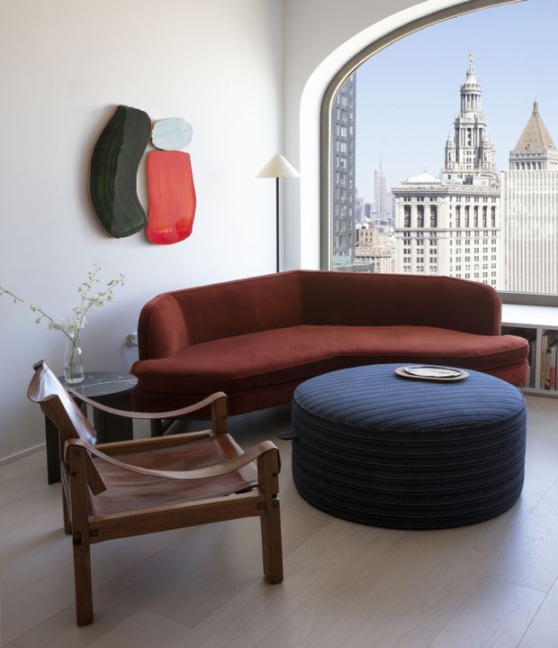 A refined high-rise living room with a curved floor-to-ceiling window overlooking a dense city skyline. The room features a deep rust-red curved sofa, a dark blue round upholstered ottoman, a wooden sling chair, a slim side table, a minimalist floor lamp, and abstract wall art. The palette is warm and sophisticated, with abundant natural light and a strong urban outlook.